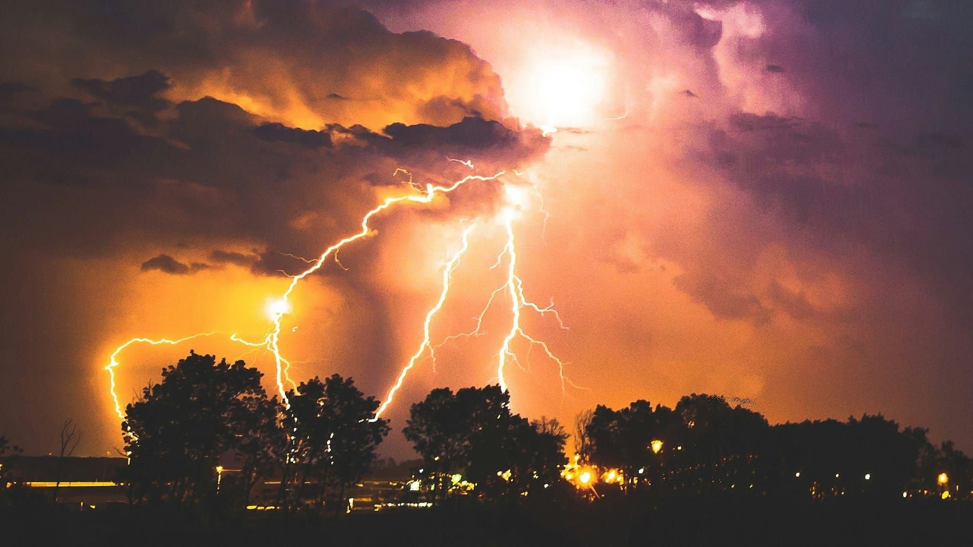 Multiple lightning bolts striking from orange and purple storm clouds over silhouetted trees and city lights at night.