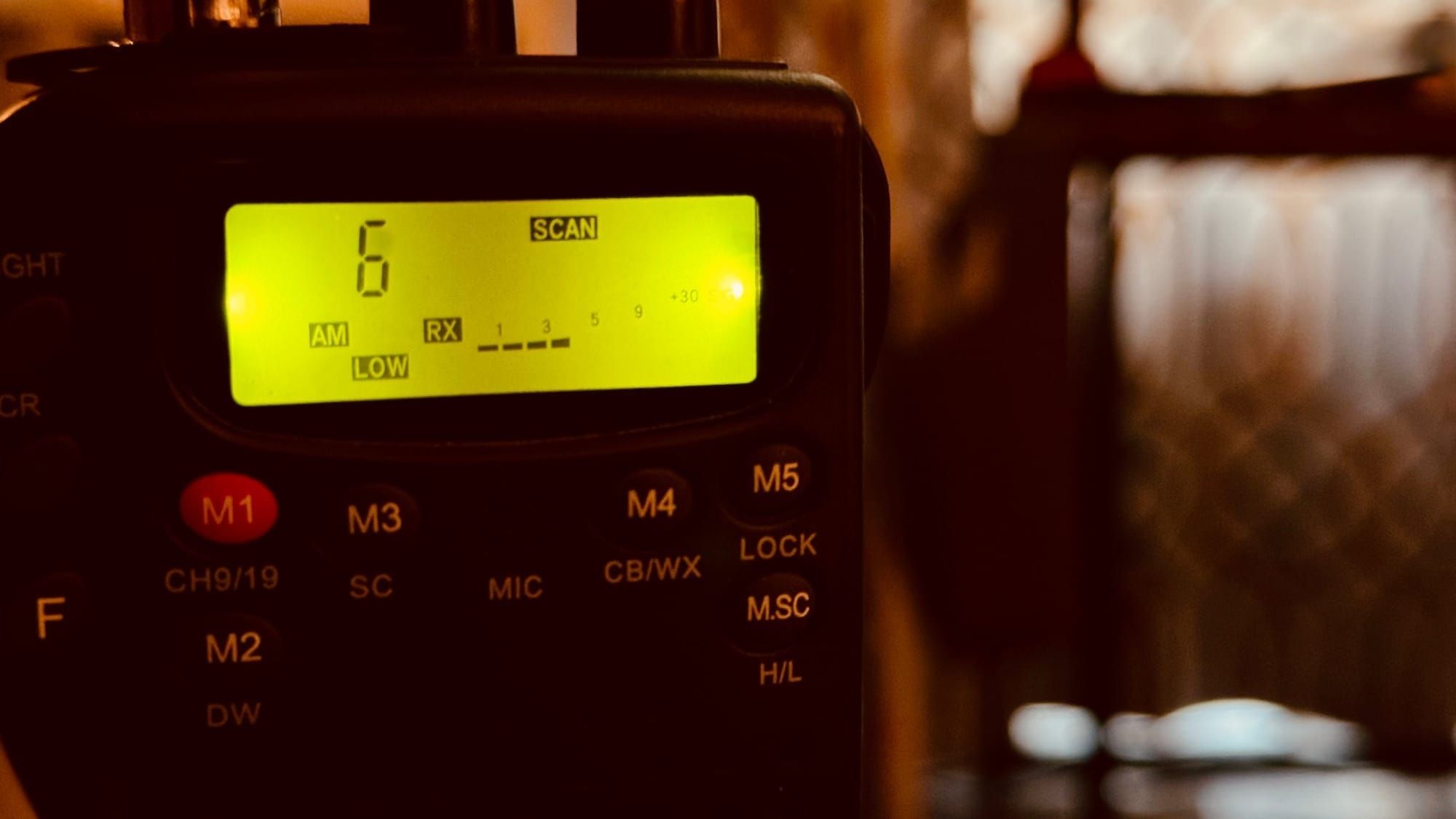 Handheld CB radio with a glowing green display showing channel 6 in scan mode, in a warmly lit room.