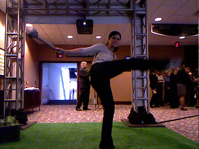 The installed booth at NAMA: truss towers, green turf underfoot, a projector overhead, and a woman mid-motion in front of the interactive screen with a crowd behind her.