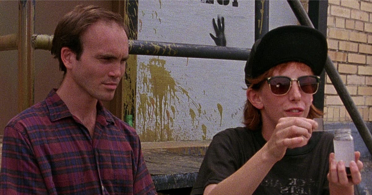 Two characters from Slacker sitting outside against a paint-splattered wall: a man in a plaid shirt looking away, and a woman in a black cap and sunglasses holding up a small glass jar.